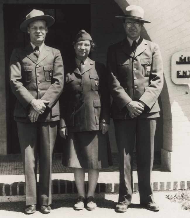 Ethel Meinzer_1942 Three uniformed rangers stand in front of a stucco building. The middle person is a woman in a skirt and flight cap while the men on either side of her wear stetson style hats. They stand with their hands clasped.