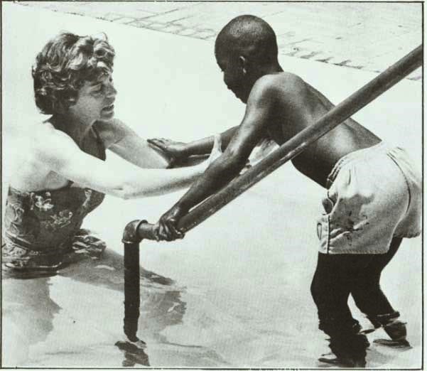Eunice Kennedy Shriver, standing in a pool wearing a swimsuit,  holds out her arms for a black child in swim trunks.