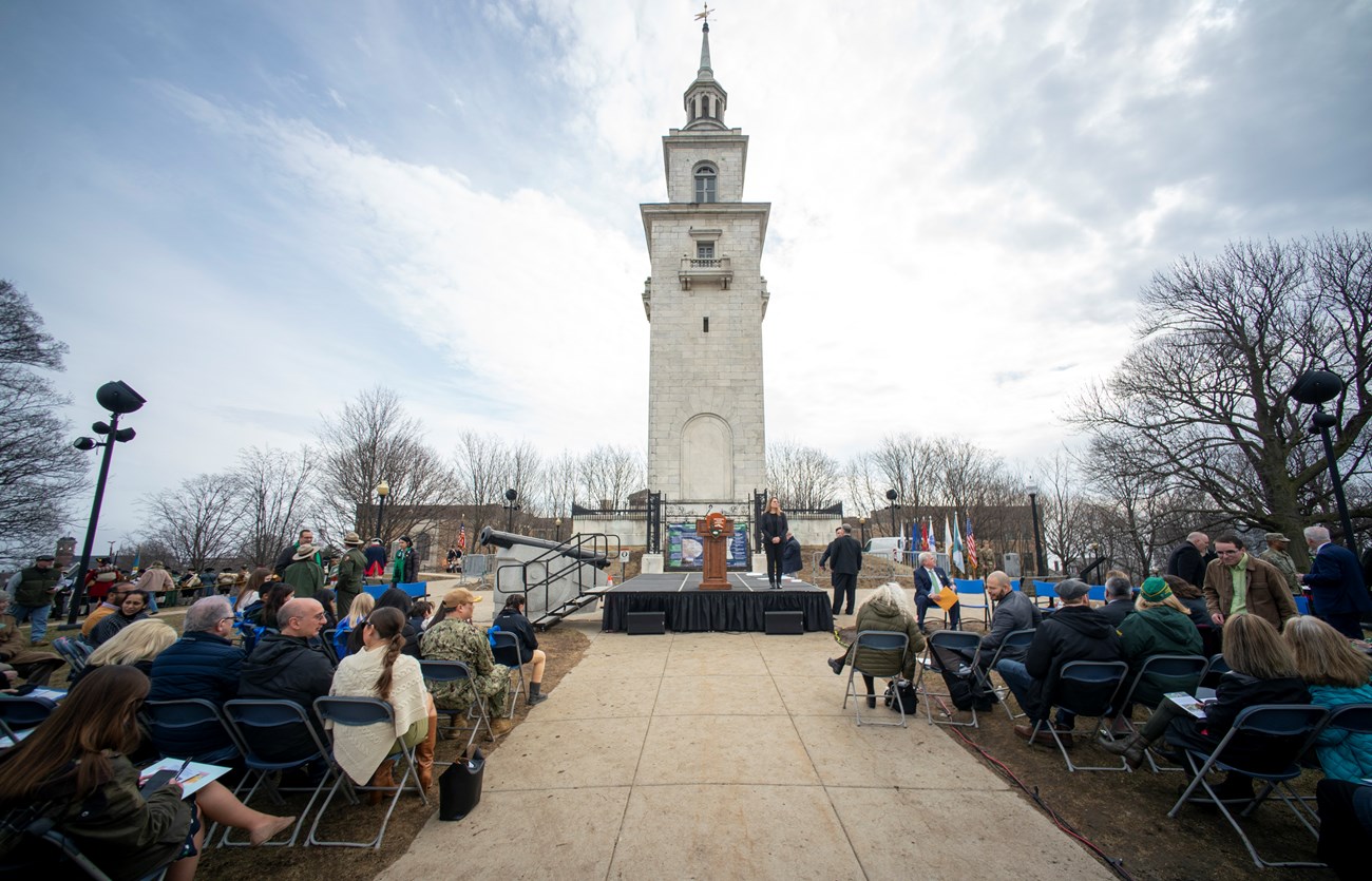Dorchester Heights Monument with stage and podium with audience sitting on either side of sidewalk leading to monument.