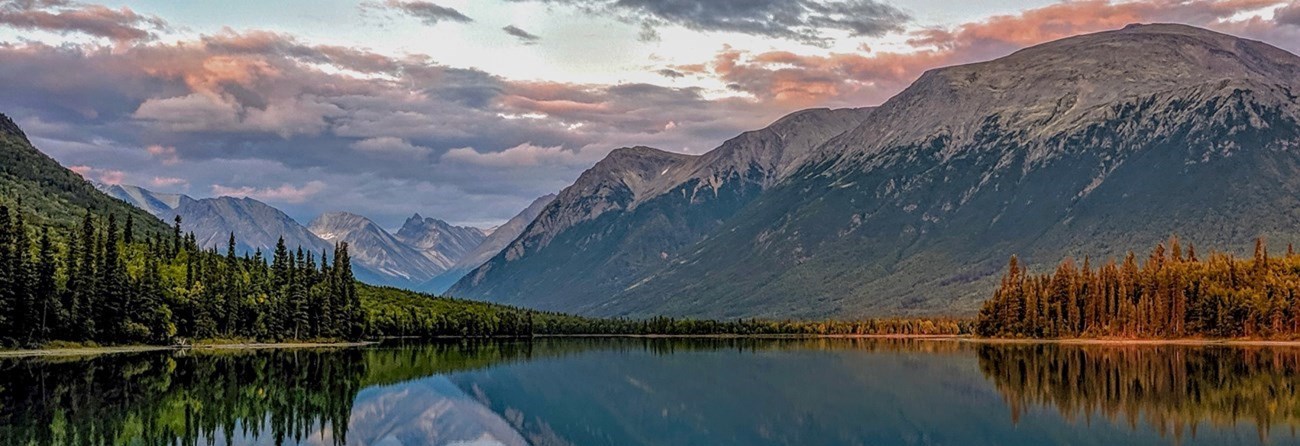 a placid lake reflects the pastel hues of an enormous mountain range at sunset.