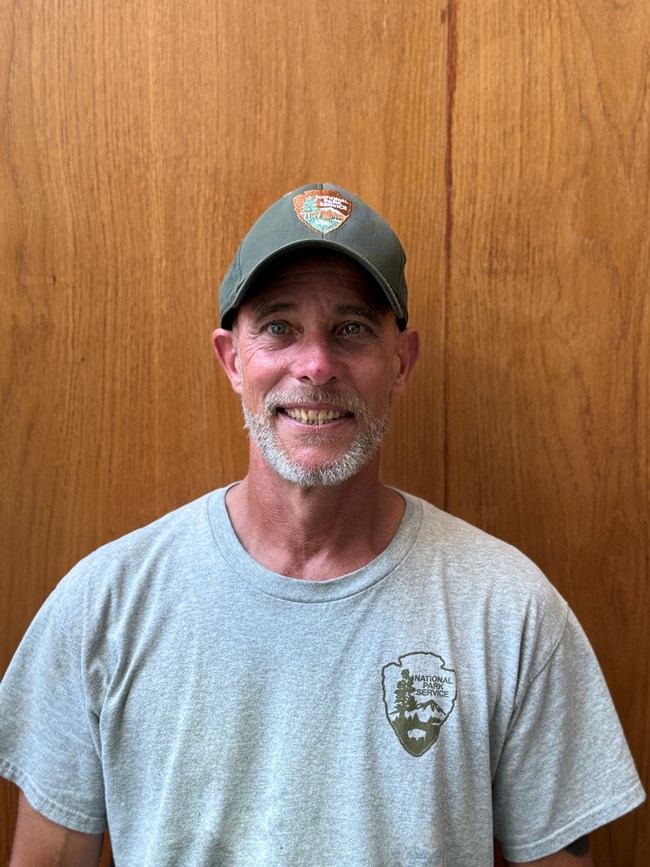 A smiling person with a gray trimmed beard, in a dark green ball cap and gray shirt.