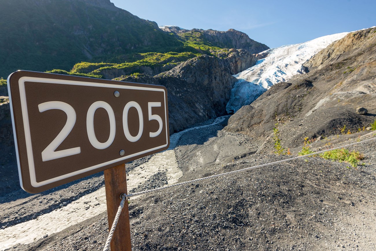 A sign reading "2005" marks the location of the toe of Exit Glacier in that year. The glacier is seen in the background.