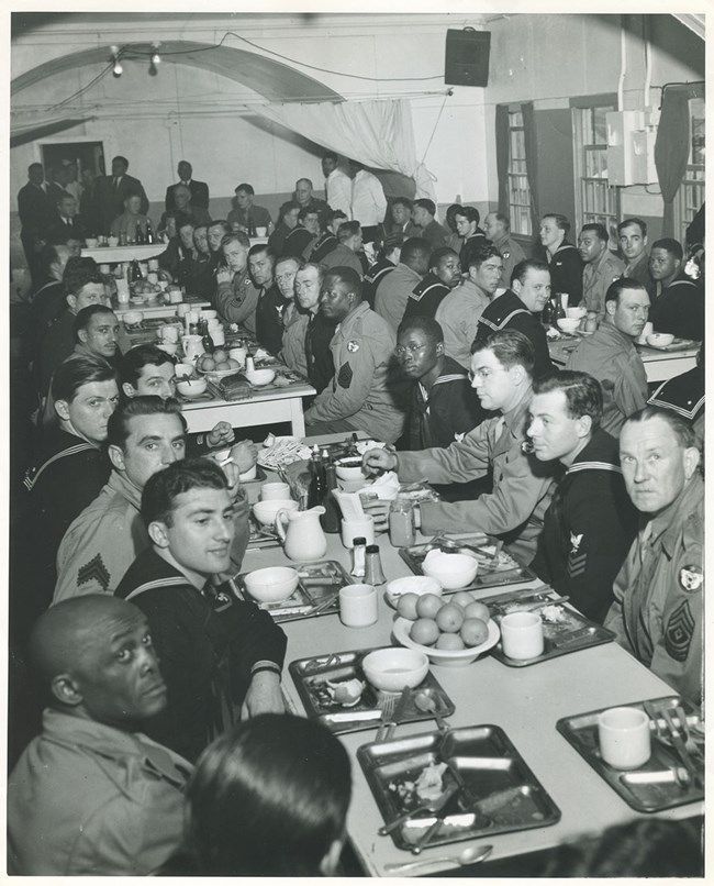 Many men in Navy, Army, and Seabee uniforms seated at long tables with trays of food.