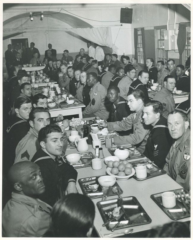 Chief Petty Officers' Mess Hall on Adak Island, with President Franklin D. Roosevelt Many men in Navy, Army, and Seabee uniforms seated at long tables with trays of food.