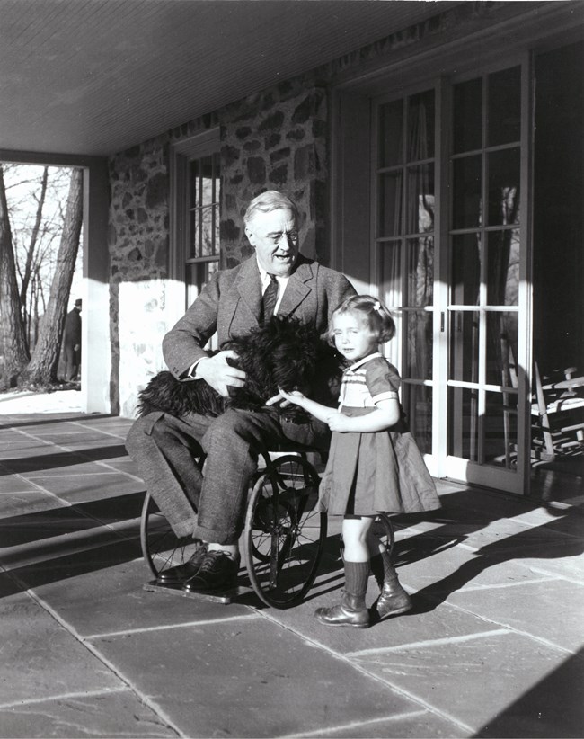 Man wearing a suit is sitting in a wheelchair on a porch. He has a black Scottish Terrier in his lap. A small female child is standing next to him as the dog licks her hand.