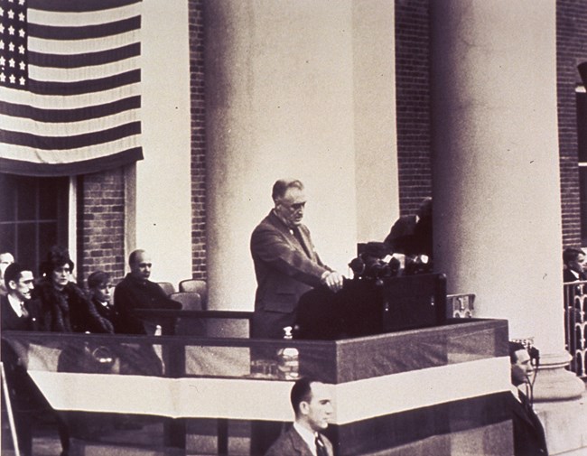 Franklin Roosevelt delivers a speech at a podium on the balcony of a columned building. People are seated behind them and a large American flag is hung in the background.