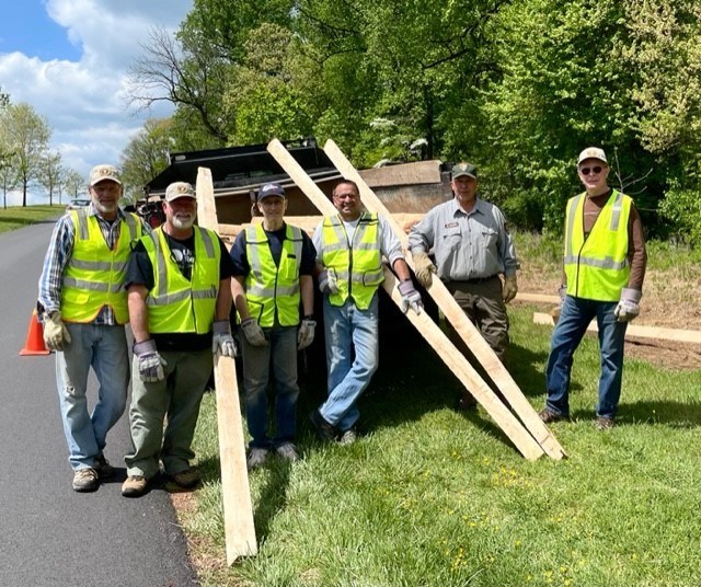 Six men wearing safety vests stand holding long timbers that are propped up on the bed of a truck.