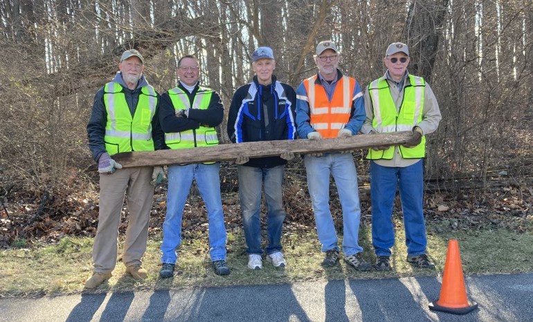 Five smiling light-skinned men wearing safety vests stand holding a timber.