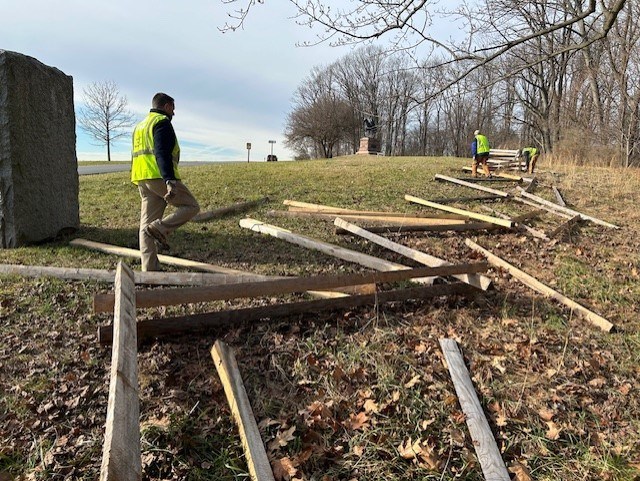 A man wearing a safety vest steps over long timbers laid out in a zig-zag pattern on the ground