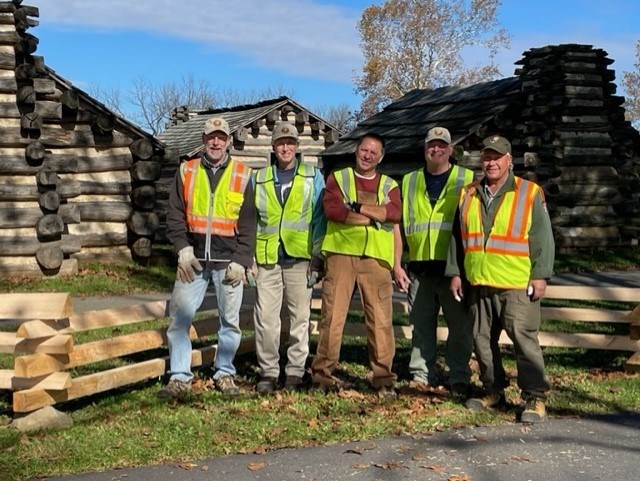 Five men in safety vests stand in front of a wooden fence with log huts in the background