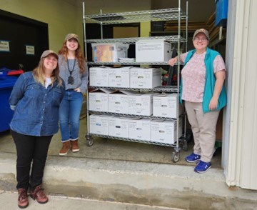 Photo of 3 people standing next to a shelf of archival boxes.