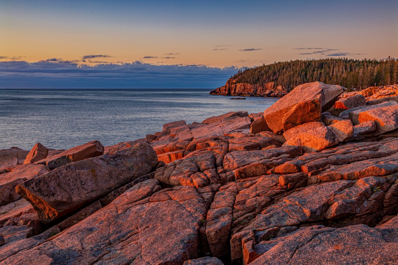 Ocean shoreline lit by low angle morning light with pink rocky boulders in foreground and calm blue water beyond