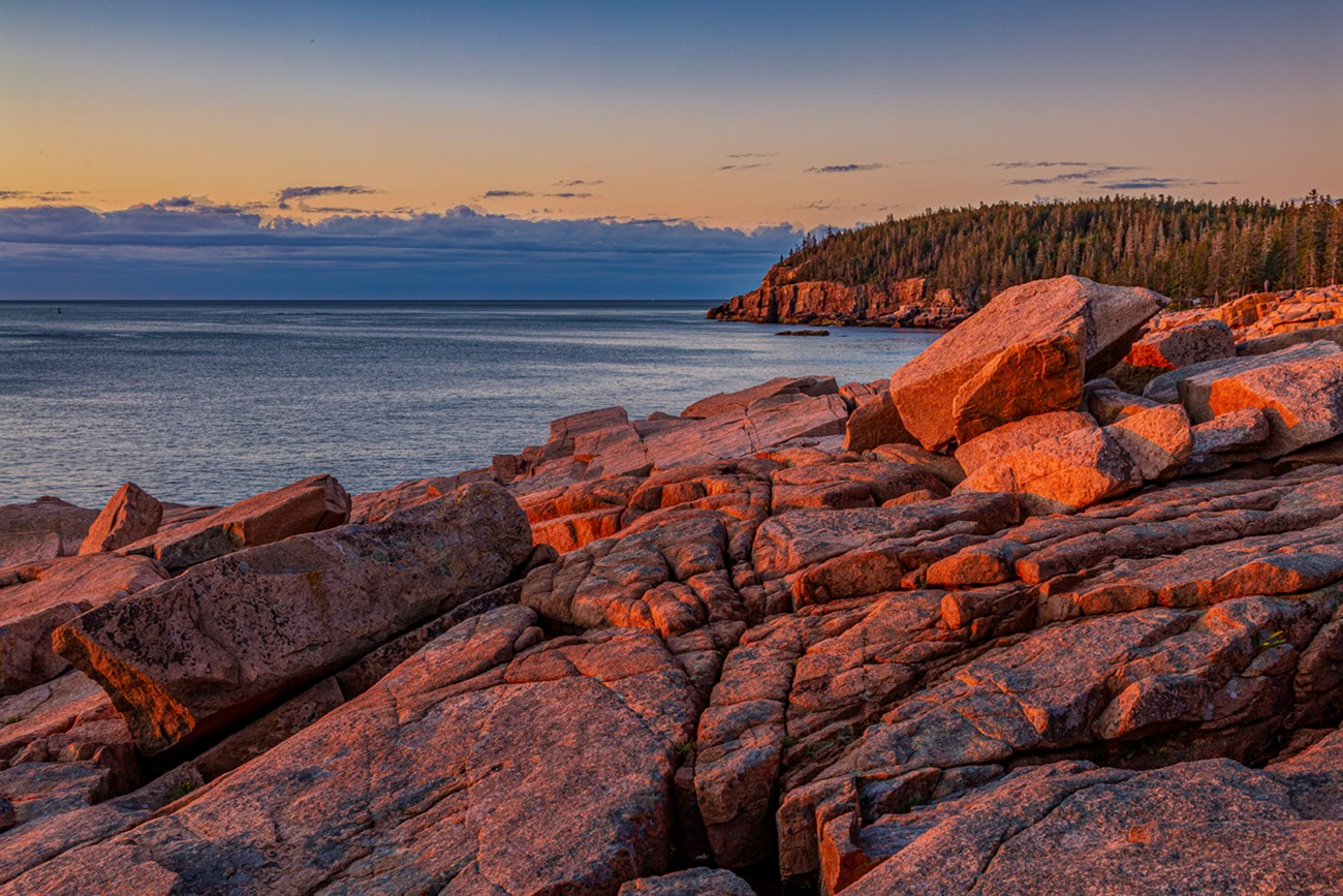 Ocean shoreline lit by low angle morning light with pink rocky boulders in foreground and calm blue water beyond Ocean shoreline lit by low angle morning light with pink rocky boulders in foreground and calm blue water beyond
