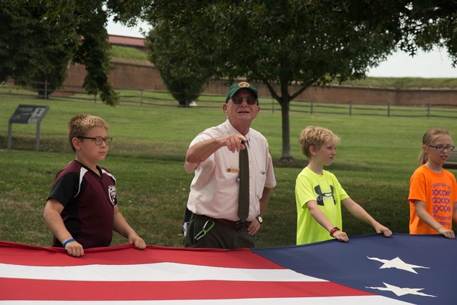 Perry Flag talk at FOMC Elderly white man, young white kids on either side of him. They are holding a very large historic replica of the American flag (war of 1812 era). Background is grass with trees.