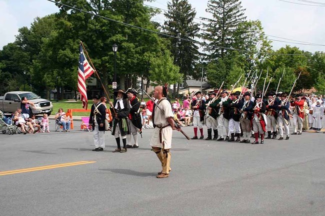 In a parade, people in period clothing and Continental Soldiers' uniform walk down a crowded, nicely decorated street.