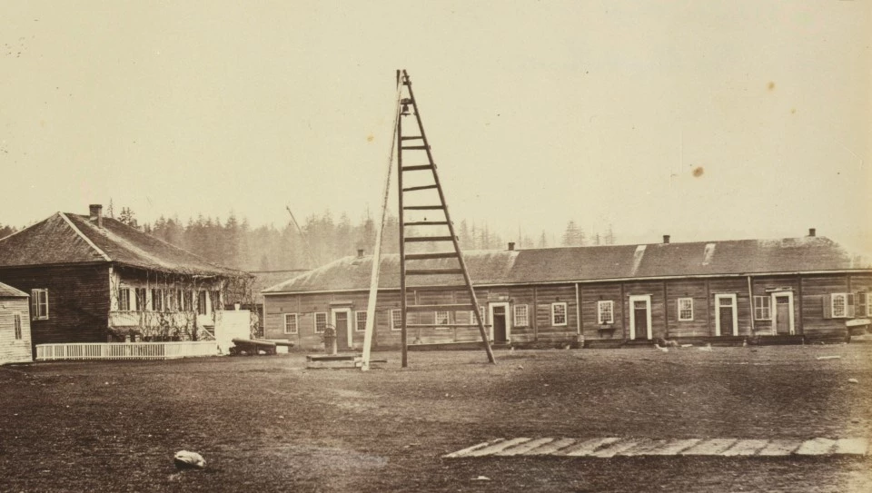 FOVA 1860 A black and white photo of 2 buildings. One is rectangular with a hipped roof, raised with a wide verandah and a short fence in front. The other building is long with at least five doors with windows arranged on each side.