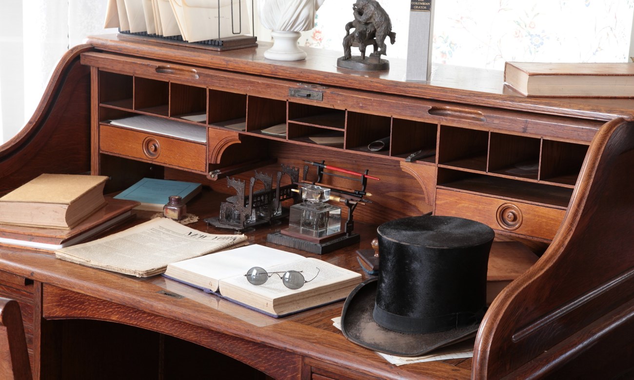 Desk covered with many items including a top hat, spectacles, books, and papers