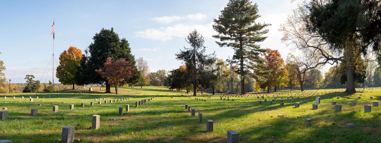 Wide view of national cemetery landscape in fall with flagpole, rows of similar headstones, and scattered trees and monuments