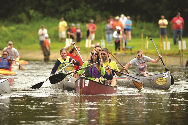 People paddling down a river in canoes with more people standing on the bank in the background.