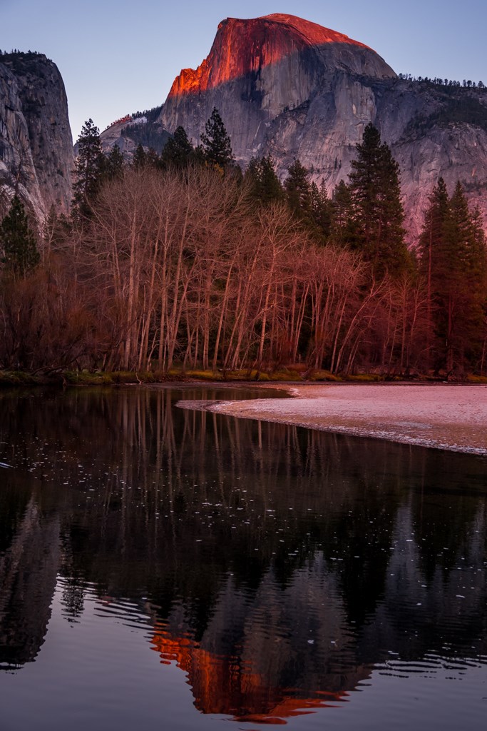 Photo of Half Dome at Yosemite National Park taken from Yosemite Valley during the Fall sunset hour. The top half of the north face of Half Dome is growing orange. Reflection on the Half Dome on the Merced River.