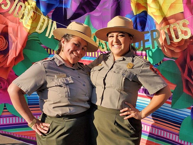 Two latina park rangers stand together in front of a colorful Dia de Los Muertos backdrop.