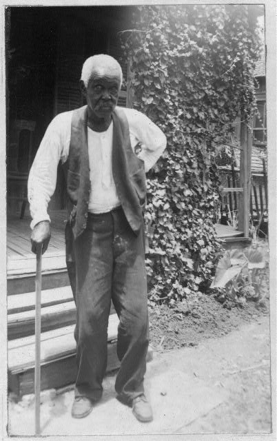 Old, African American man stands outside his front porch, supporting himself with a cane. He wears dark pants and a dark vest with a white shirt underneath.