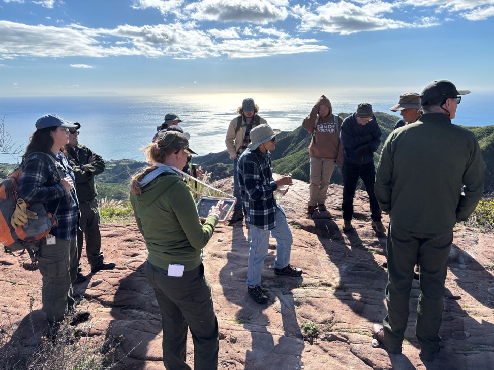 Eleven people dressed for cool weather stand on a bare rock outcrop overlooking the sea on a mostly clear day.