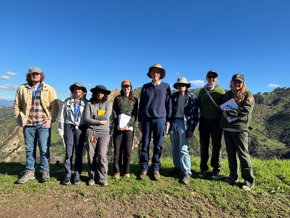 Eight people dressed for cool weather stand on a grassy ridge in a line facing the camera.