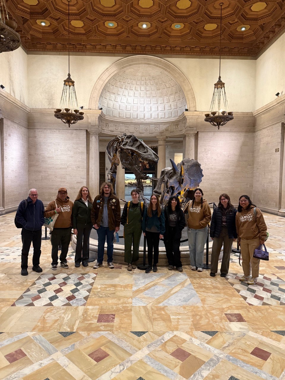 Ten people stand in a line facing the camera in front of a skeletal mount of a predatory dinosaur confronting a three-horned dinosaur in a large room.