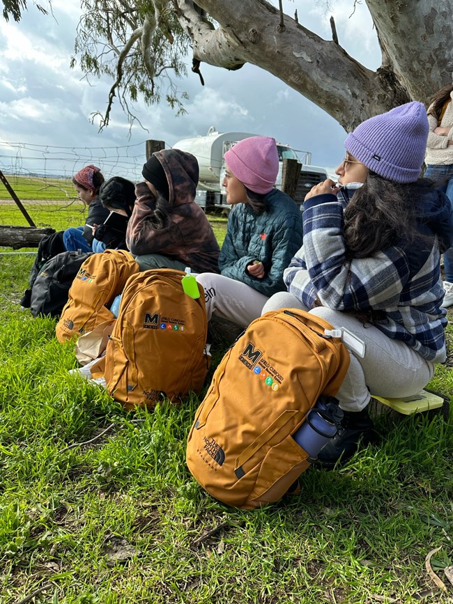 Five young students participating in a field science event sit on a low bench under a tree sitting with new backpacks and wearing warm clothes.