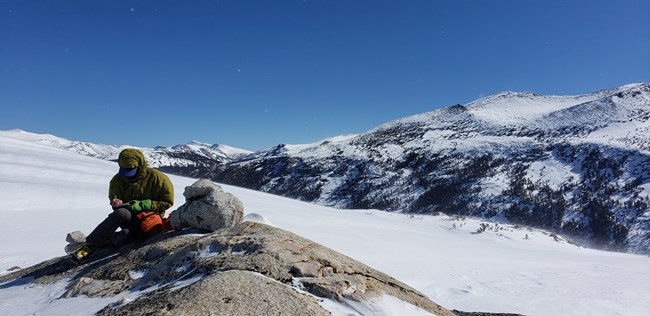 Scientist in warm jacket and gloves sits on rock surrounded by snow in a high mountain landscape.