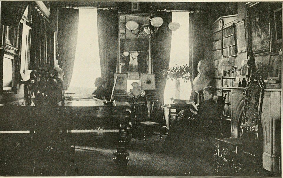Two women seated in room with several bookshelves, piano, and fireplace