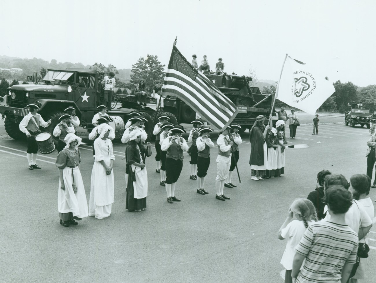 A group of reenactors standing in rows with some playing fife and drum. An American Flag and Bicentennial flag are flown.