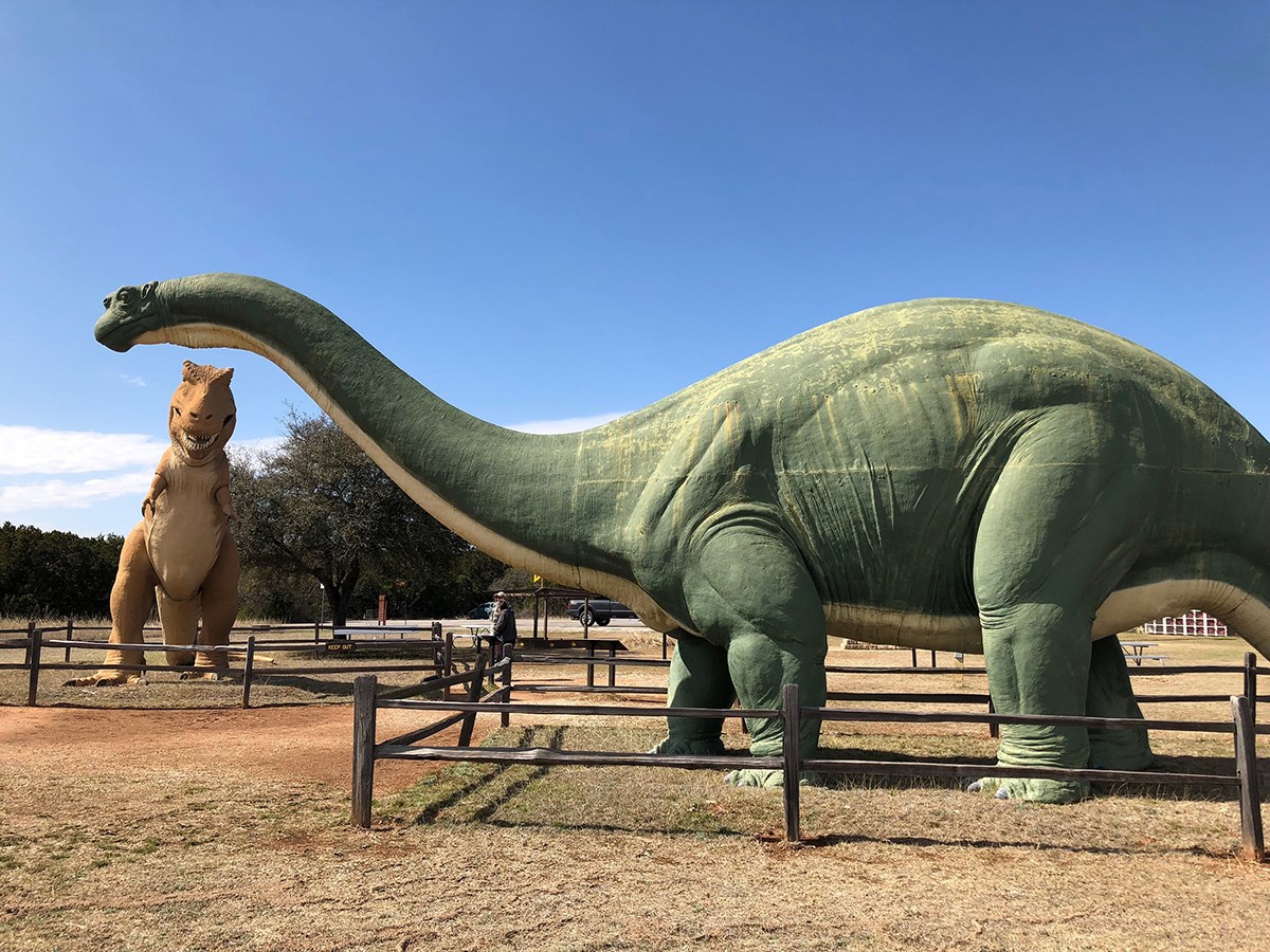 Statues - brontosaurs in foreground T-rex in background. Rail fence surrounds each statue.