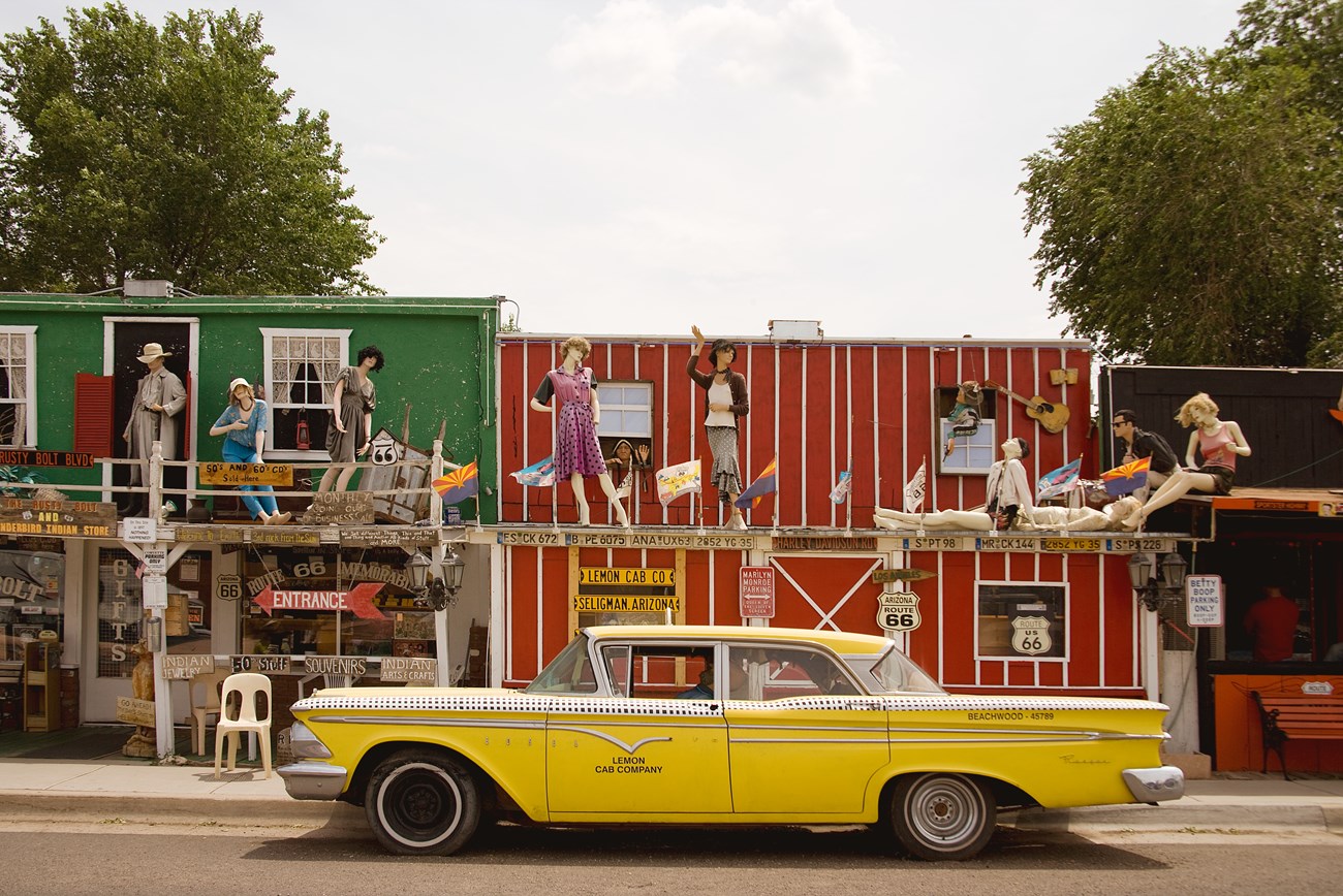 8 mannequins stand on a makeshift store front balcony with an old yellow 4 door car parked in front.