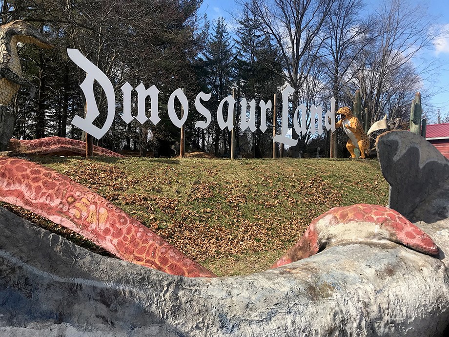 Letter sign reads Dinosaur land – Dinosaur statues on both ends and in the lawn in front of sign.