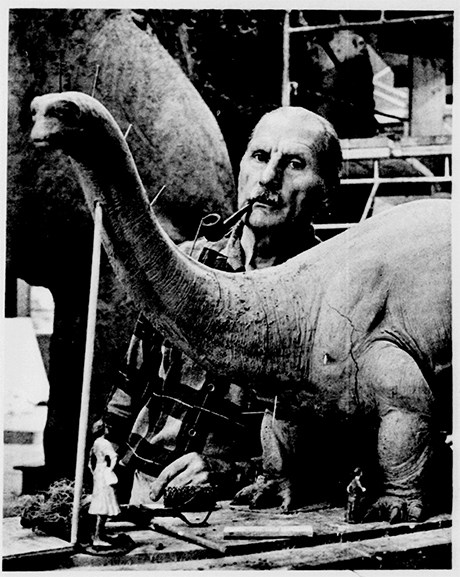 Balding man with a smoking a pipe behind a model of a long neck dinosaur.