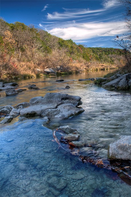 Slow flowing river around gray rocks, brown and green trees on the shore, and wispy white clouds in a blue sky.