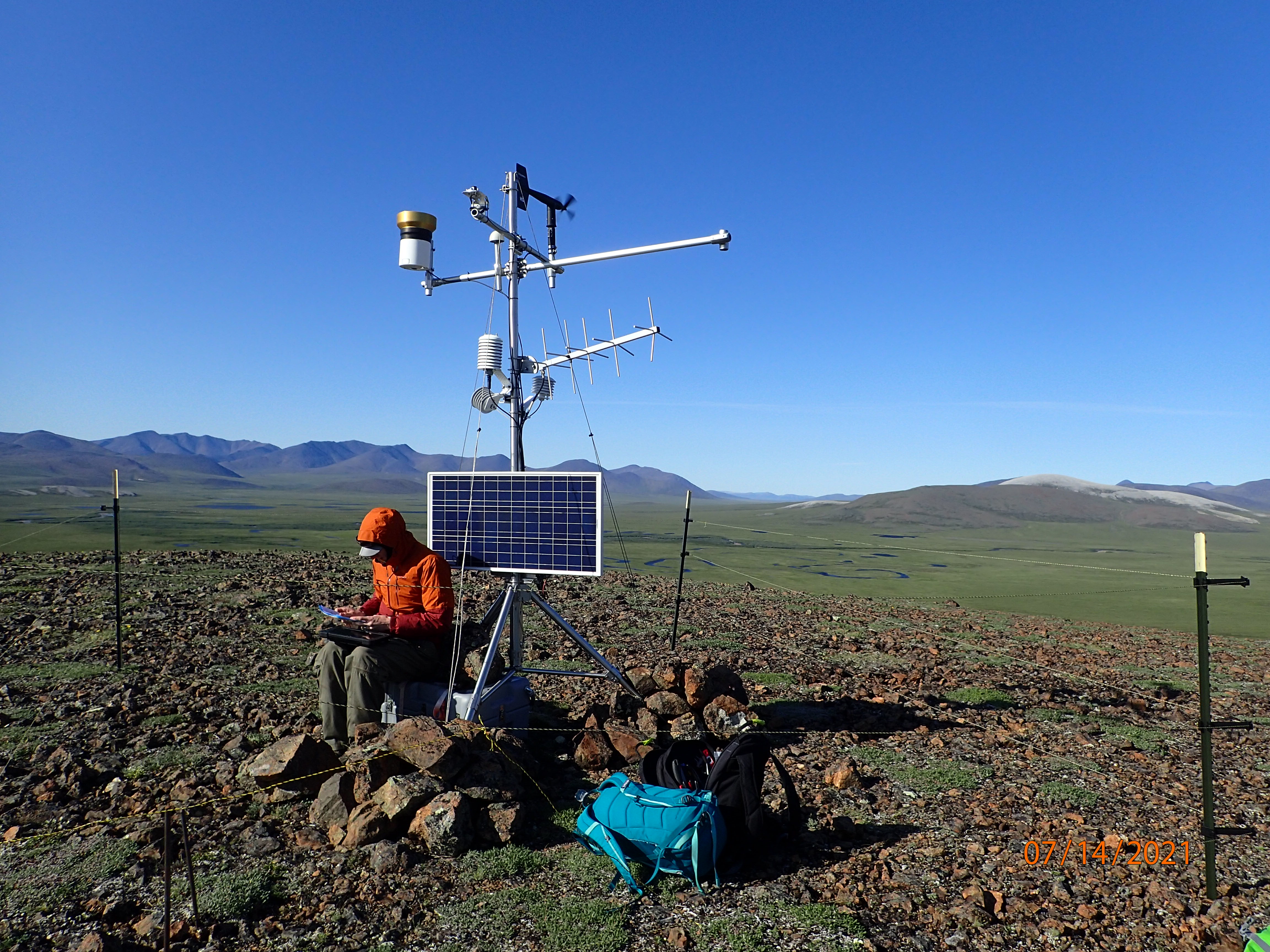 A person works on a climate station in an open expanse of tundra.
