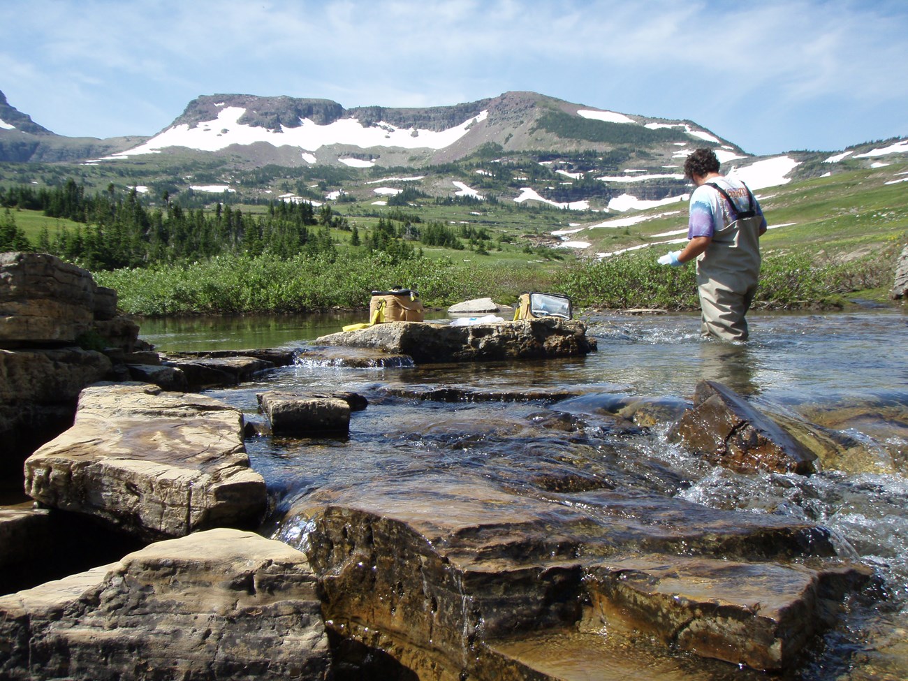 Technician collects water from an alpine stream with snowy mountains in background.