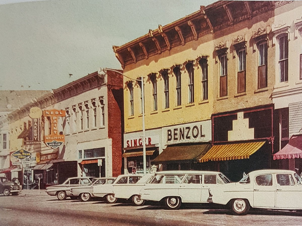City street with brick building store fronts and 1950s era cars and station wagons parked in front.