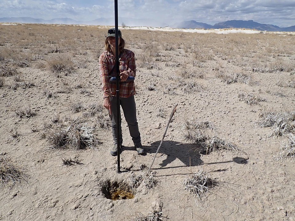 EC-30 Spring A person next to an upright pole pointing to a depression in a flat, white, sandy area with dispersed clumps of grass.