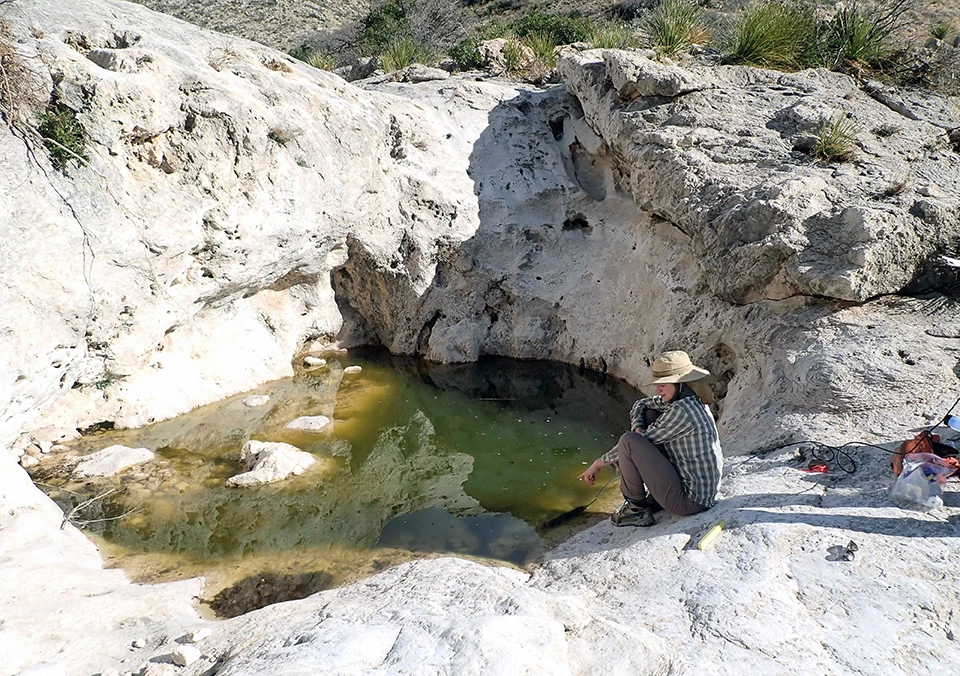Slaughter Pot Hole A green-tinged pool in a bedrock basin with a crouched crew member at the edge of the pool pointing at the edge of the water.