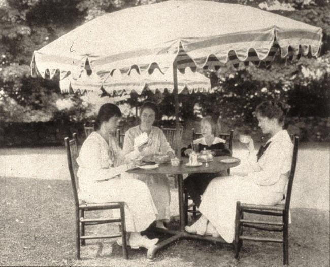 Four women sit at a table under a large umbrella drinking tea.