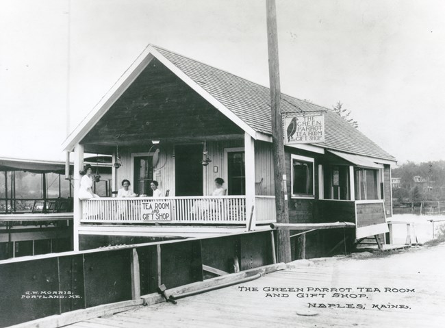 Four women sit on the porch of a small house on a dock with a sign reading the green parrot tea room and gift shop.