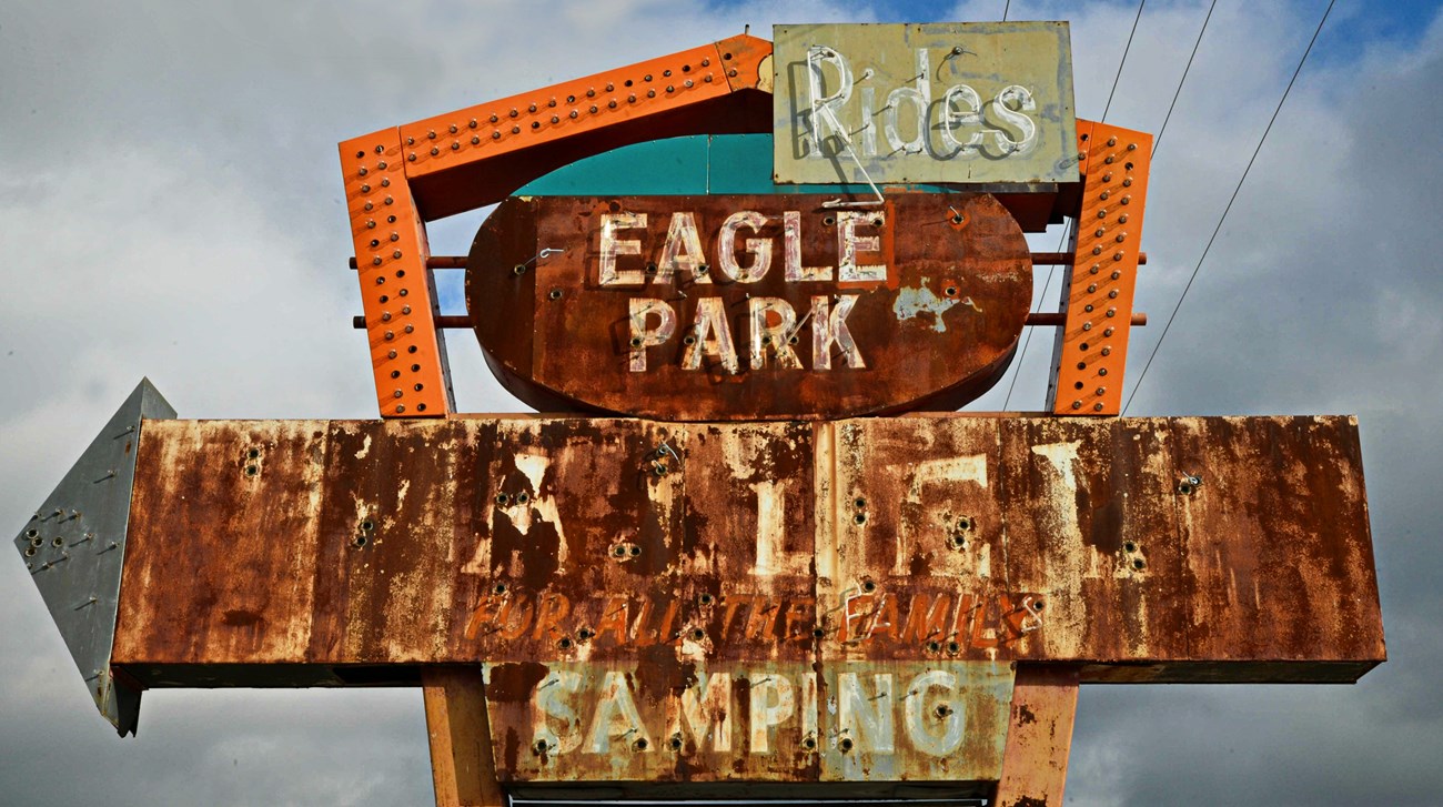Rusting sign with arrow missing light bulbs with text reading rides, Eagle Park, and other lettering not legible.