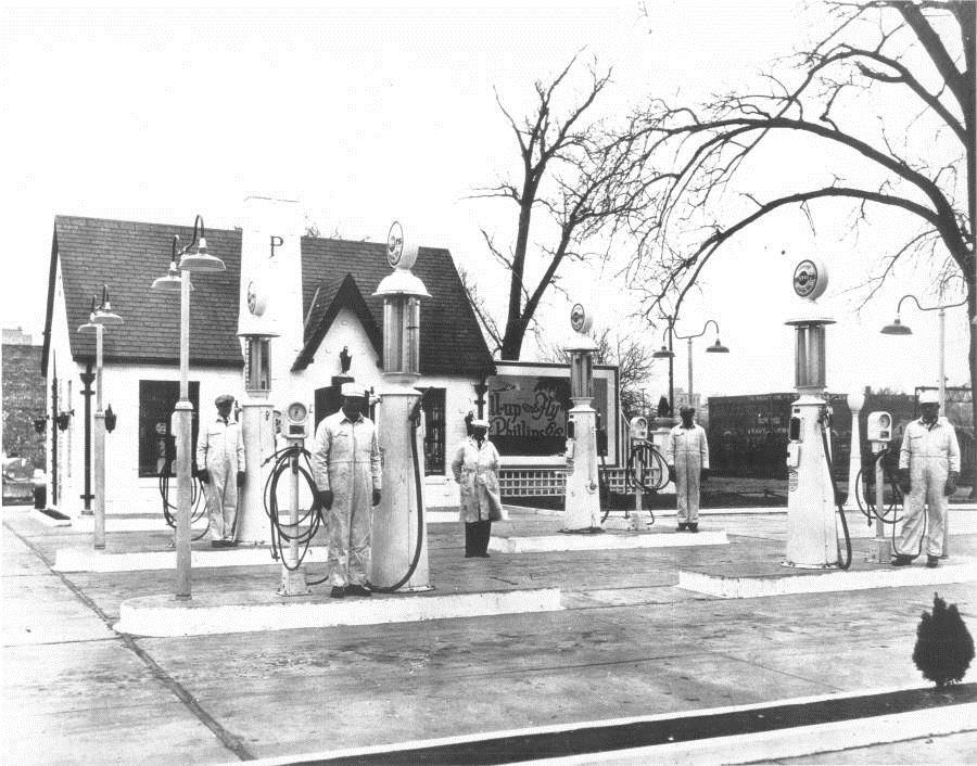 5 men in white coveralls stand next to white gas pumps with a small white brick building in background.