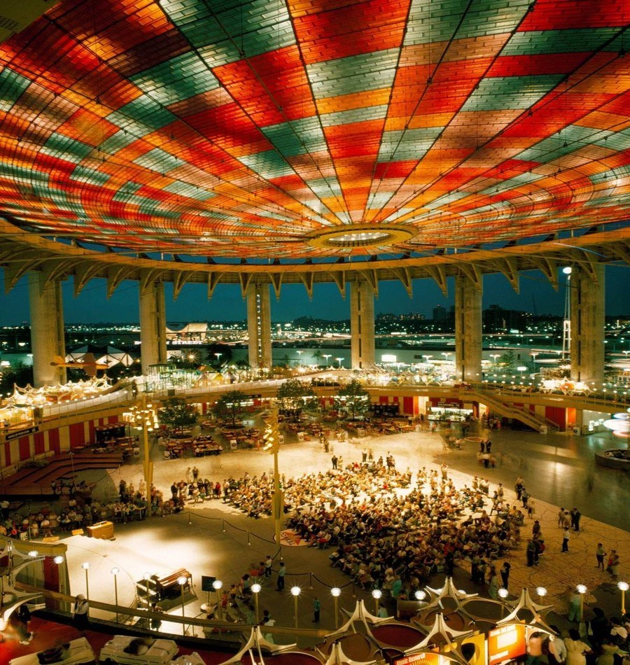 Looking down: large crowd in a 2-acre open air round pavilion; ceiling random mosaic red and green rectangles.
