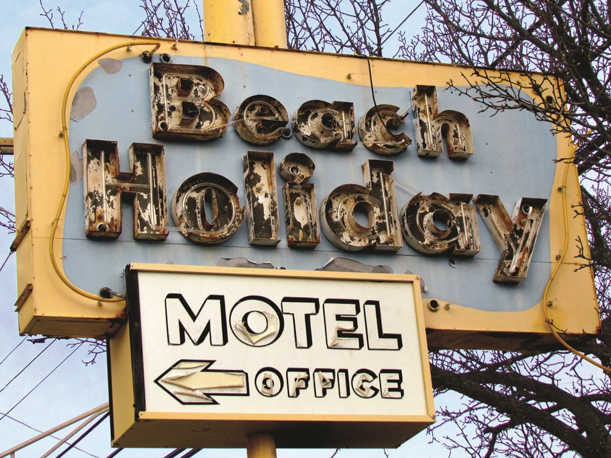 Weathered neon sign with text Beach Holiday Motel Office. An arrow points left.