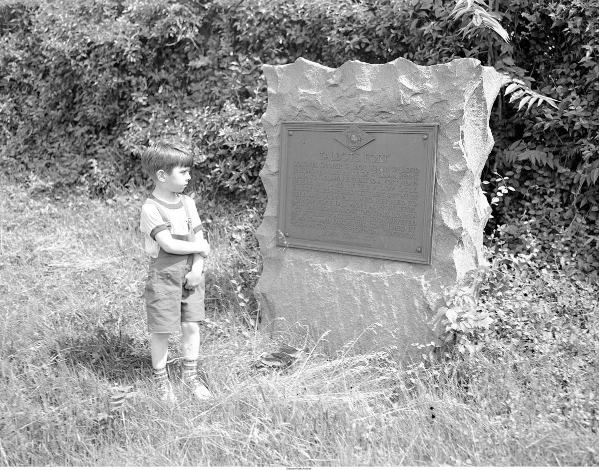 Young boy wearing overall shorts stands next to a large stone with historical plaque about Talbot’s Fort.